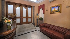 A lobby with a reception desk, a brown leather sofa, a staircase, and a framed painting on the wall. - Hotel Piccolo di Corduneanu Aurora (Termini Imerese)