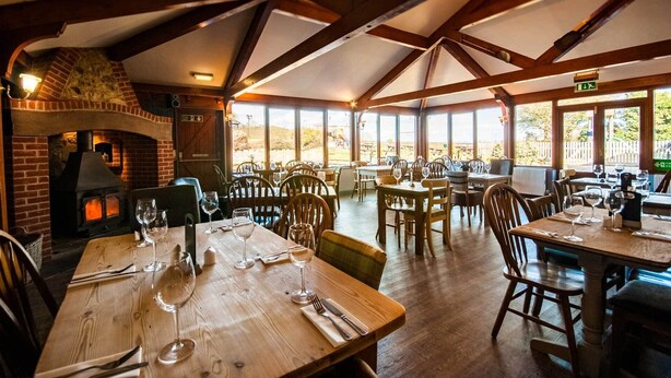 A restaurant with wooden tables and chairs, a brick fireplace, and large windows offering a view of the outdoors. - The Chequers Inn (Ventnor)
