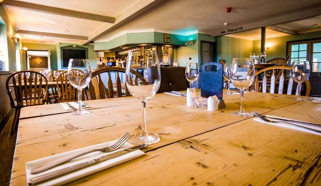 Un restaurant avec des tables en bois disposées pour le service, un bar à l’arrière-plan et un panneau indiquant l’emplacement des toilettes.