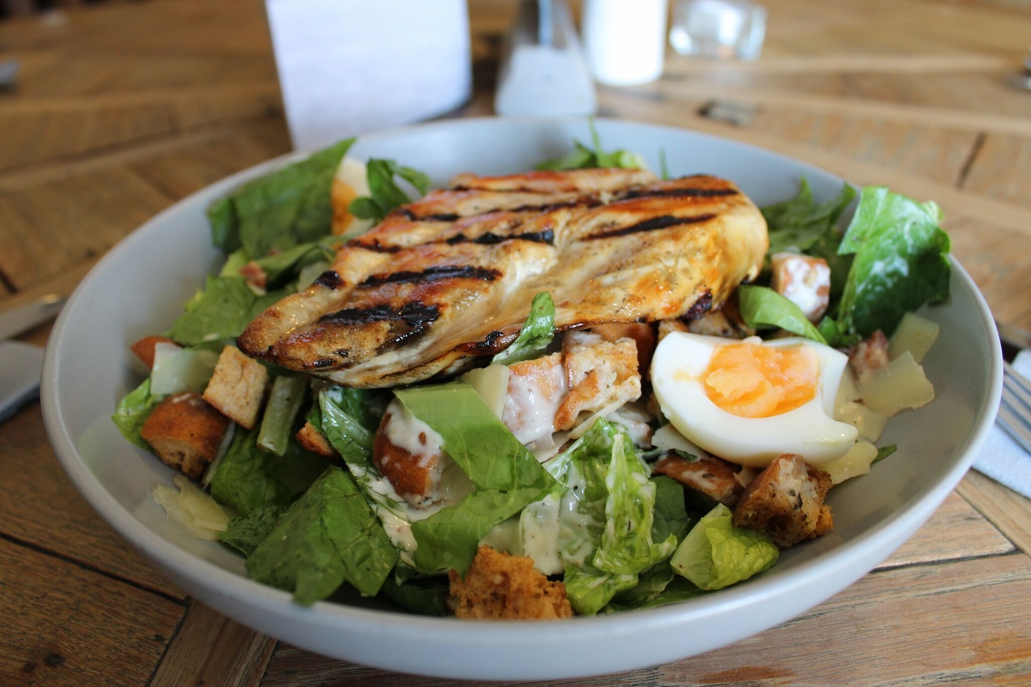 Een saladekom met gegrilde kip, een gekookt ei en croutons op een houten tafel.