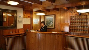 A wooden reception desk with a receptionist, framed pictures on the walls, and a shelf with items. - Hotel Escalar (Panticosa)