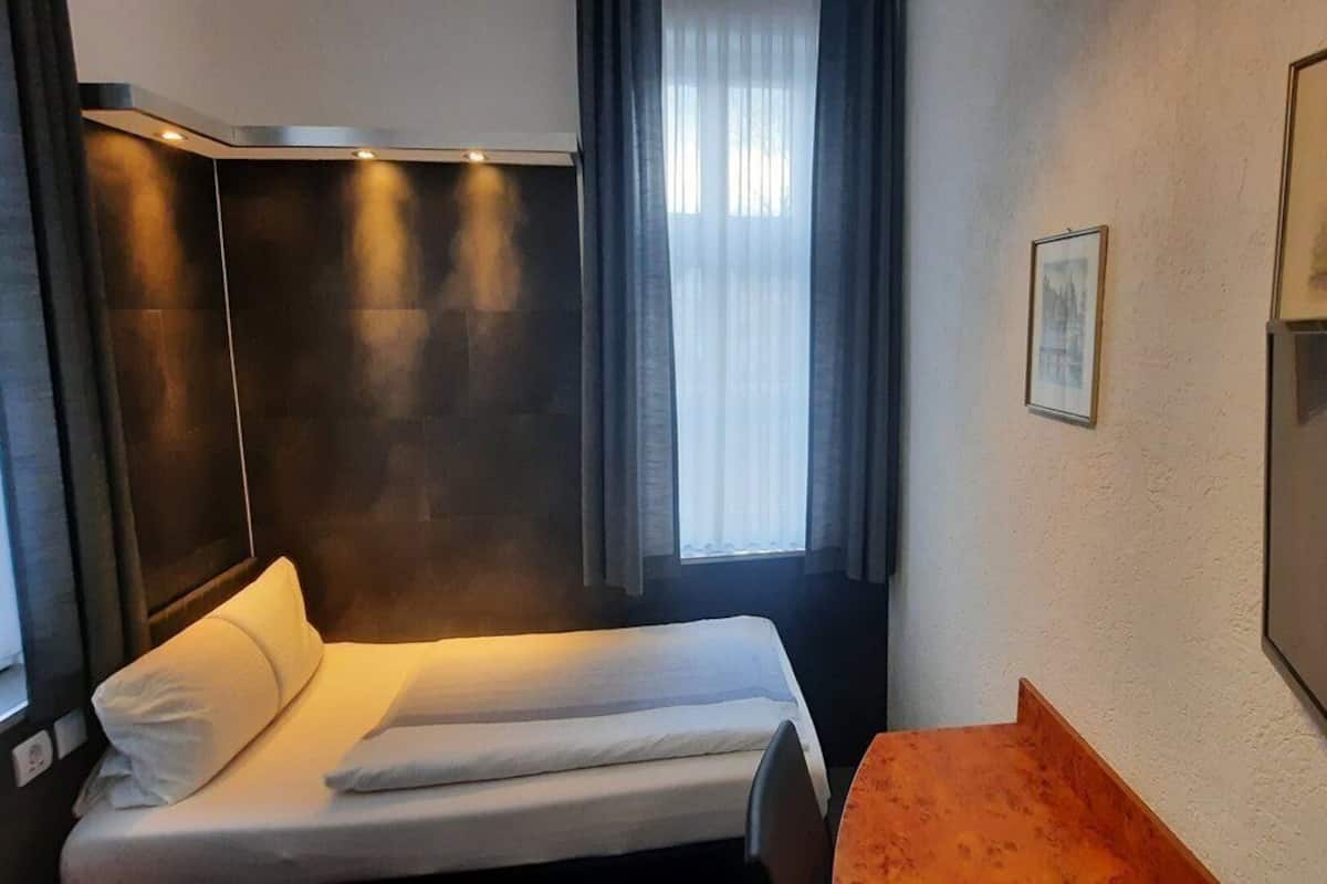 A small, compact hotel room with a single bed, a wooden table, a television, and a window with curtains.