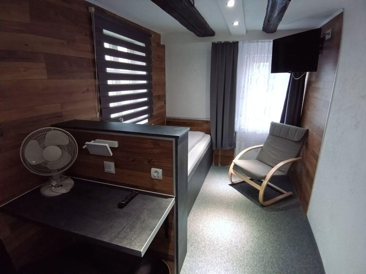 Habitación de hotel compacta con cama, escritorio con ventilador, silla y televisor montado en la pared.