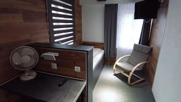 Habitación de hotel compacta con cama, escritorio con ventilador, silla y televisor montado en la pared.