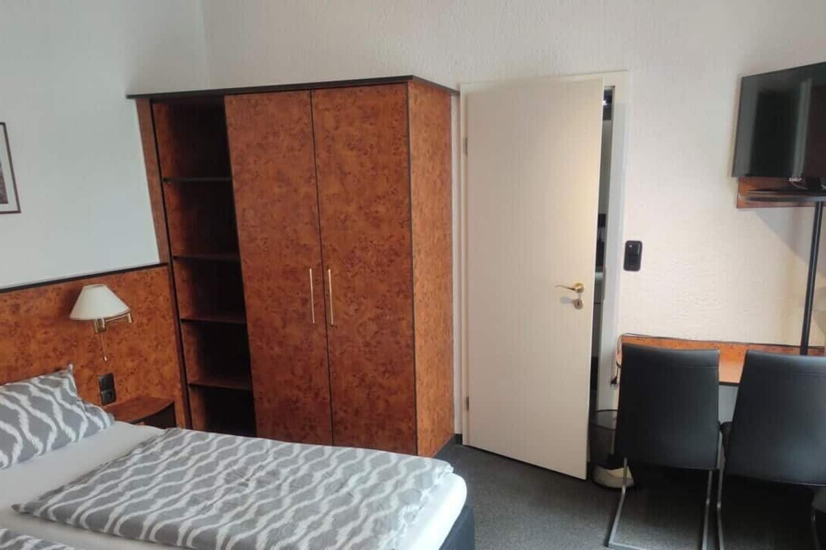 A hotel room with a bed, a wardrobe, a desk with chairs, a television, and a door.