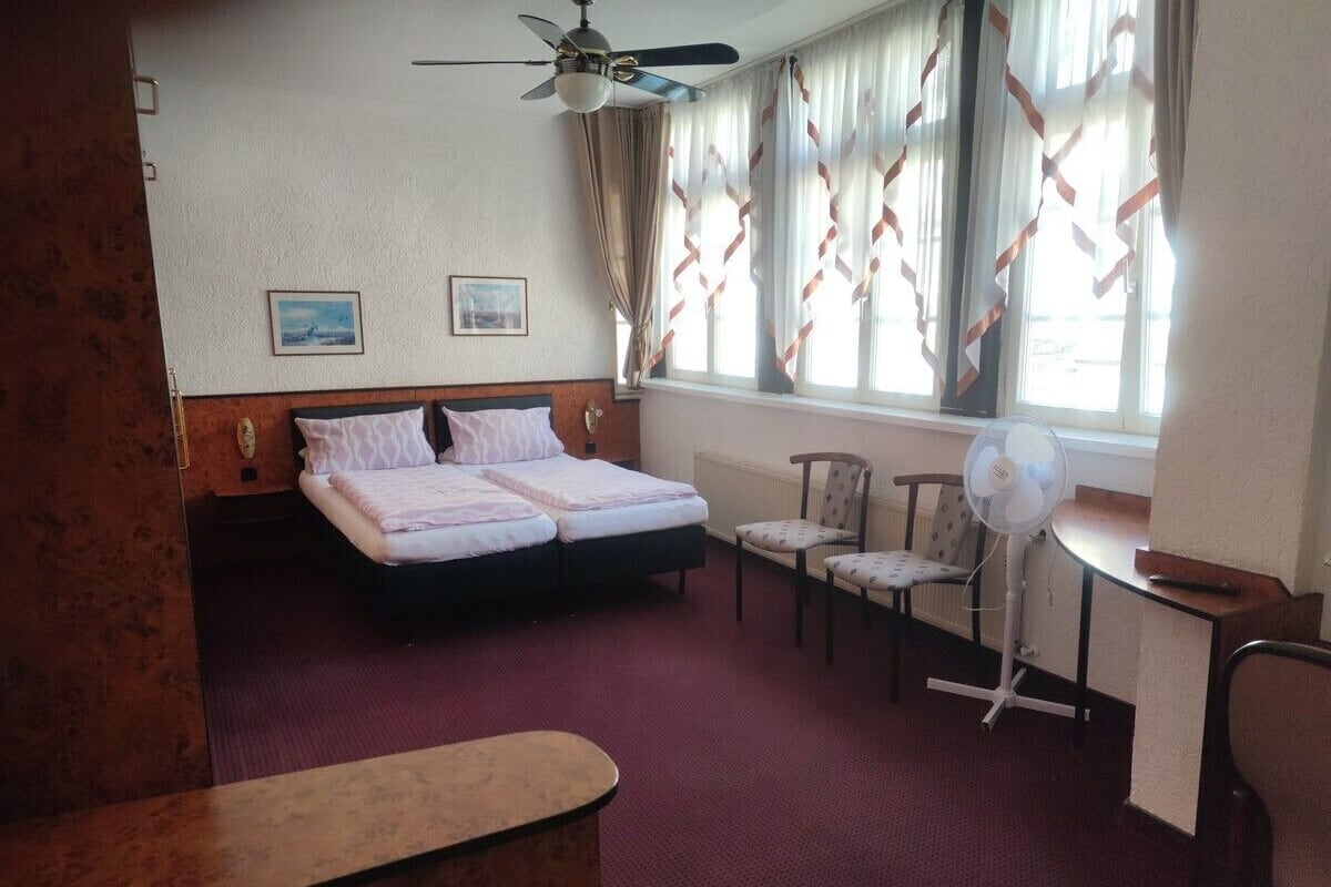 A hotel room with a double bed, two chairs, a desk, a fan, and a ceiling fan.