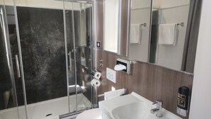 A bathroom with a glass-enclosed shower, a white sink, a mirror, and a towel dispenser. - Hotel Corveyer Hof (Hoexter)