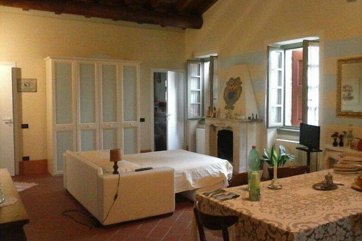 A room with a bed, a table with a patterned tablecloth, a chair, a fireplace, and a door leading to another room.