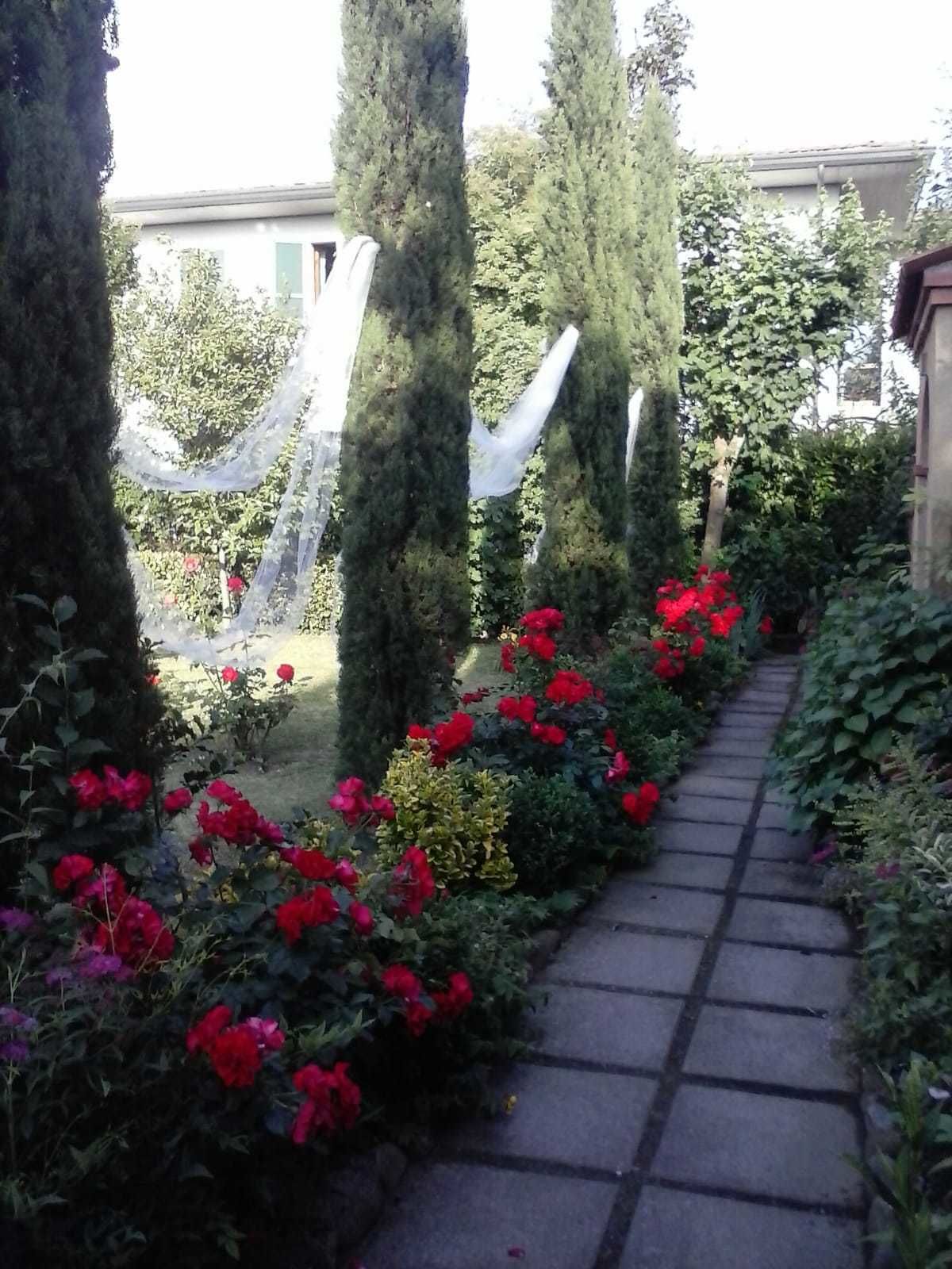 Un sentier de jardin bordé de fleurs rouges et d’arbres imposants, qui mène à un bâtiment à la façade blanche.
