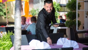A waiter serving a table at an outdoor cafe with wicker chairs and a red and white checkered tablecloth.