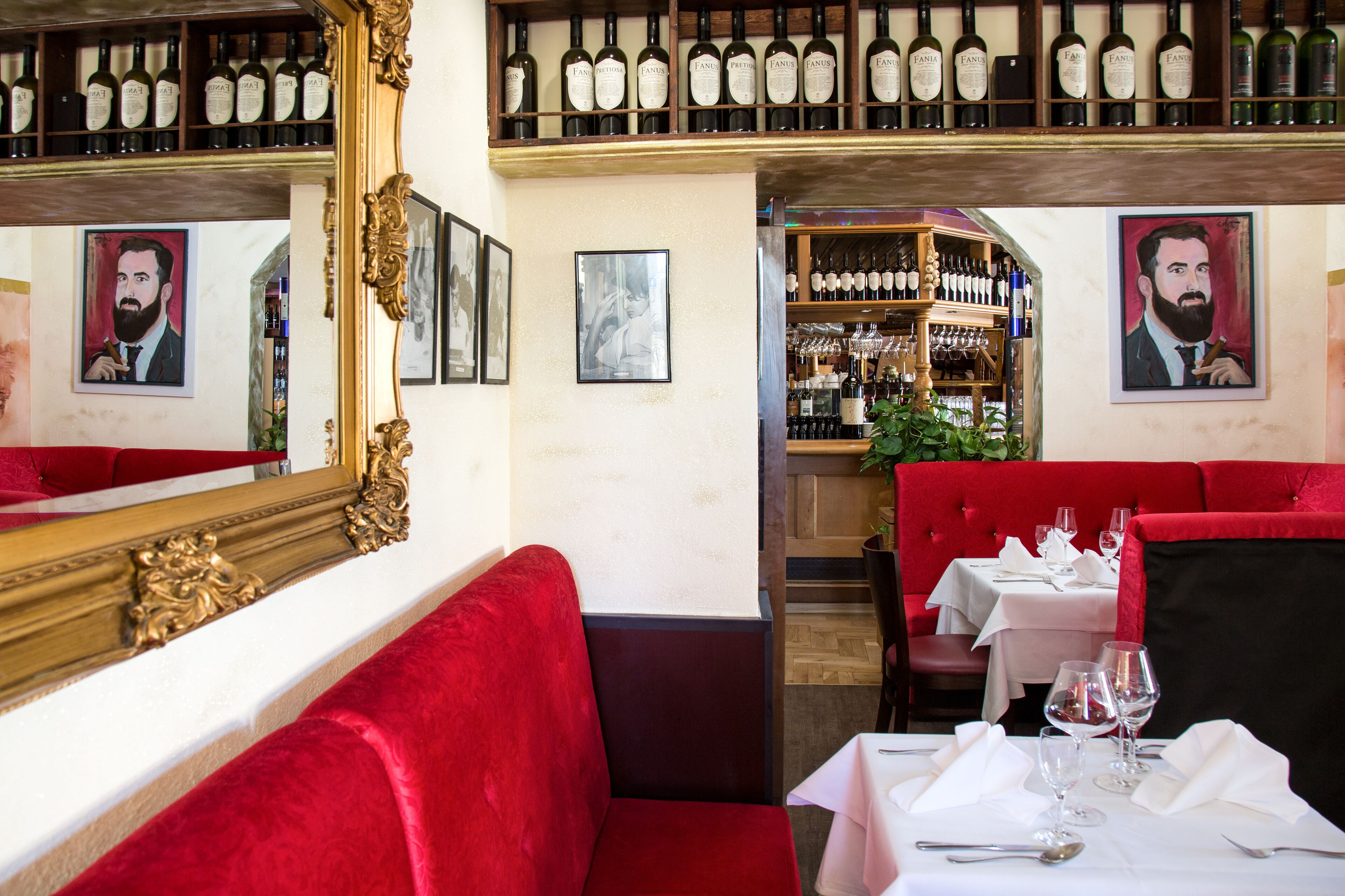 L’intérieur d’un restaurant présente des sièges rembourrés de couleur rouge, un présentoir à bouteilles de vin au-dessus du bar et des portraits encadrés accrochés aux murs.