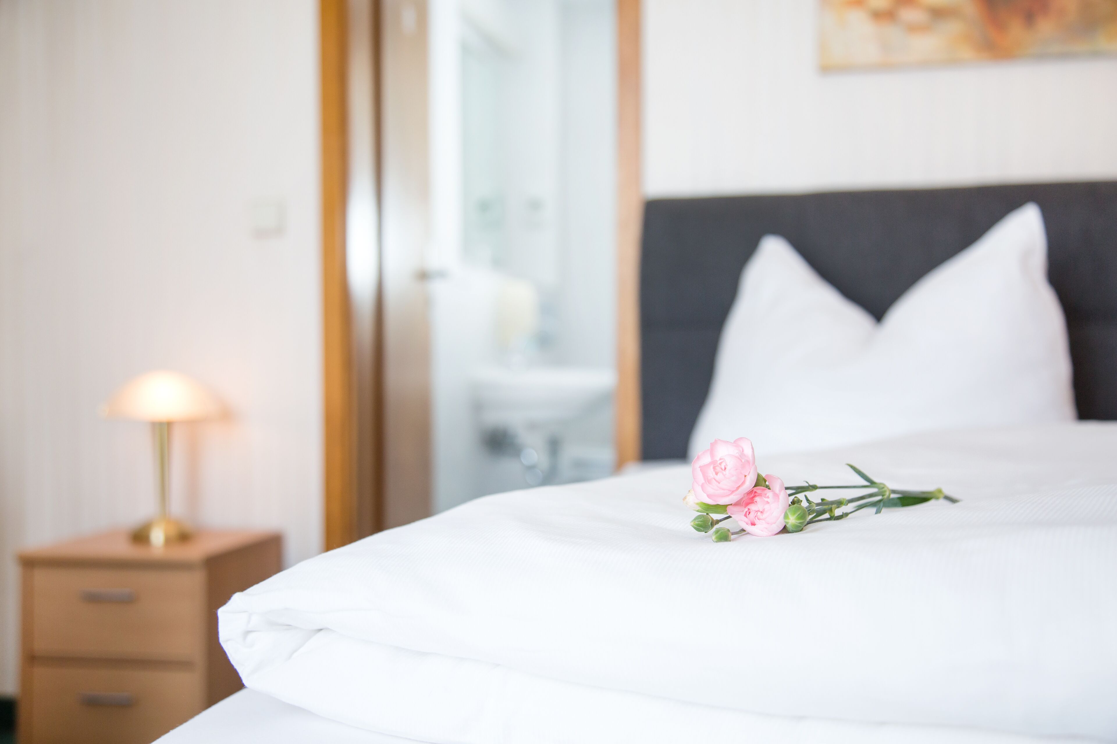 Una cama bien hecha con sábanas blancas, una lámpara de noche y una flor rosa sobre la almohada.