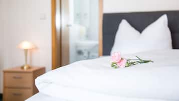 Una cama bien hecha con sábanas blancas, una lámpara de noche y una flor rosa sobre la almohada.