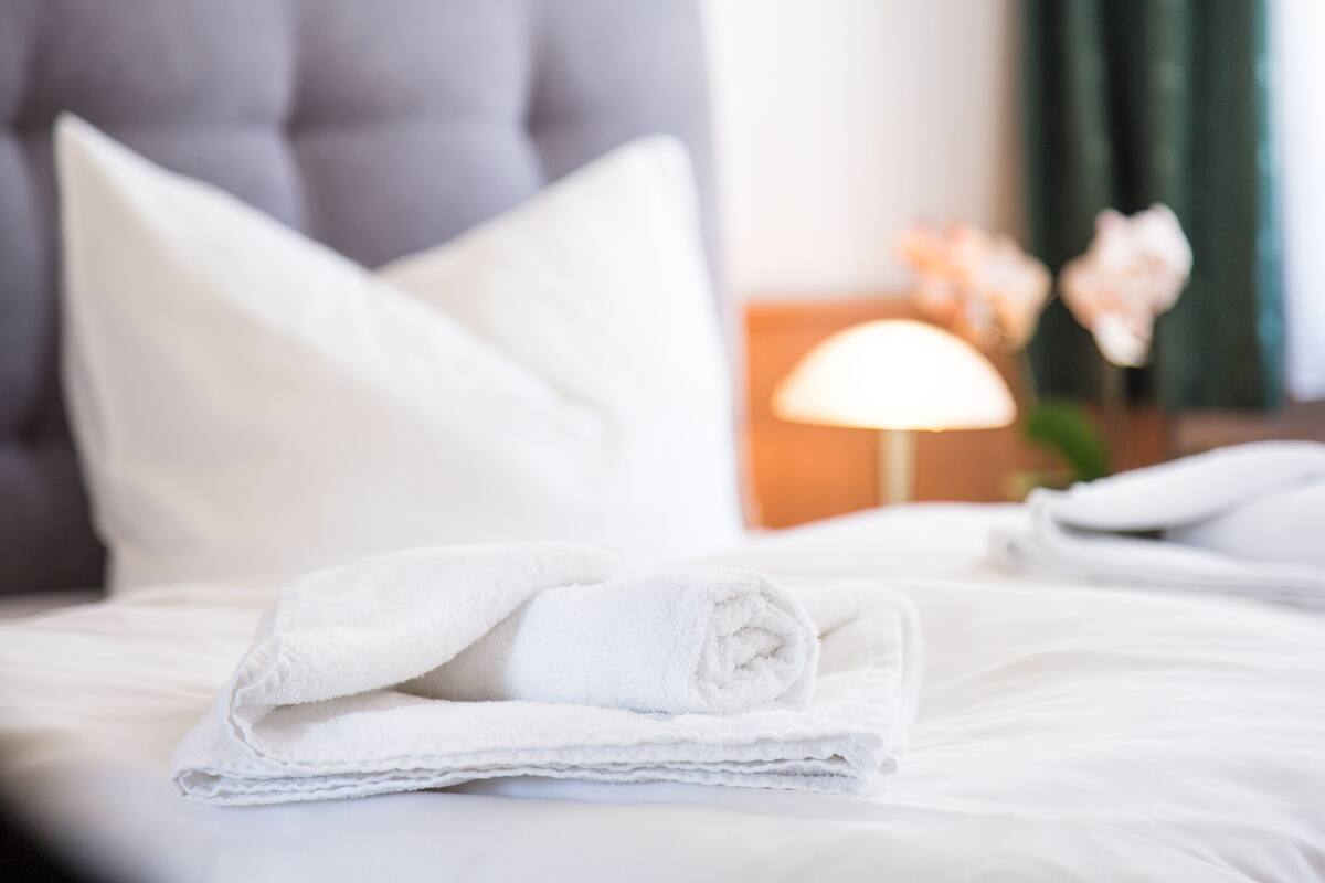 Una cama bien hecha con sábanas y almohadas blancas, una lámpara sobre una mesita de noche y un jarrón con flores al fondo.