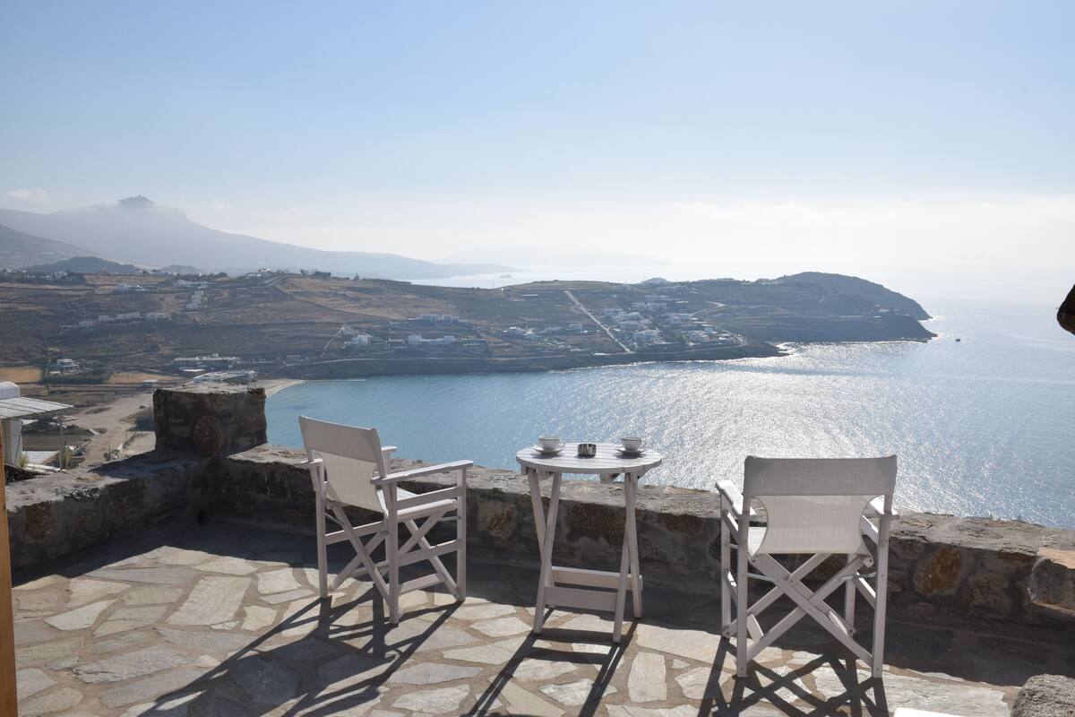 Eine Terrasse mit Steinboden, zwei weißen Stühlen und einem kleinen runden Tisch mit Tassen und Untertasse.
