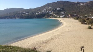 A coastal landscape with a sandy beach, a clear blue sea, and a hillside with white buildings. - Andromeda Mykonos Villas & Suites (Mykonos)