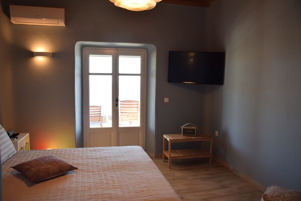 A bedroom with a bed, a small wooden table, a television mounted on the wall, and a door leading to an outdoor area. - Andromeda Mykonos Villas & Suites (Mykonos)
