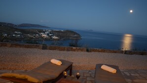 A seaside lounge with two sunbeds and a table, overlooking a coastal town and the sea. - Andromeda Mykonos Villas & Suites (Mykonos)