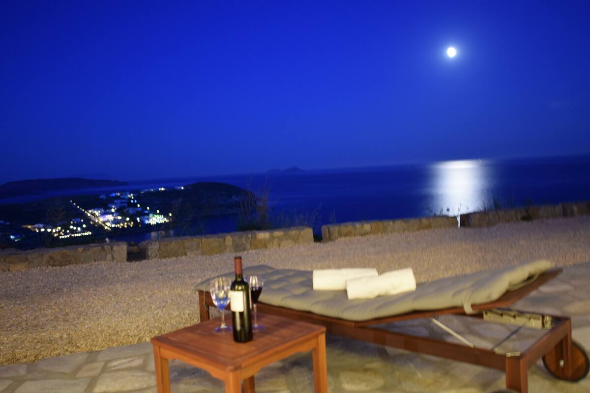 Ein Liegestuhl und ein Tisch mit einer Flasche und einem Glas auf einer Terrasse mit Blick auf die Küste in der Nacht.