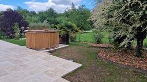 Ein Holzbott im Freien auf einer Steinterrasse, umgeben von einem angelegten Garten mit Bäumen und Sträuchern.