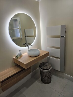 A modern bathroom with a round mirror, a white sink, a wooden vanity, and a wicker waste basket. - Provencal house with swimming pool in magnificent wooded park in the heart of Périgord (Ribérac)