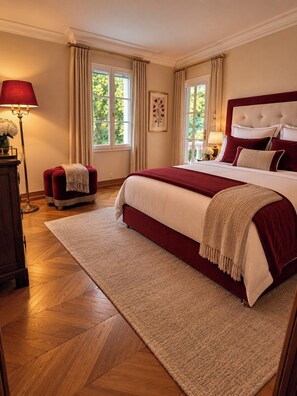 A bedroom with a large bed, a nightstand, a lamp, a window with curtains, and a wooden floor.