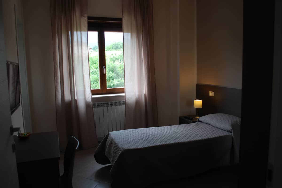 A hotel room with a bed, a nightstand with a lamp, a window with curtains, and a radiator.