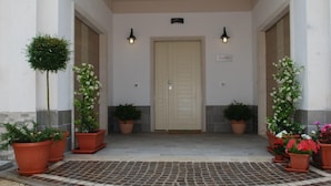 Entrada de um edifício com portas duplas, plantas em vasos e duas luzes na parede.