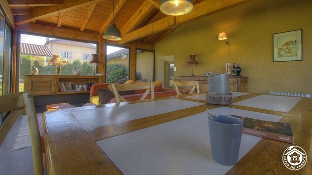 Um quarto acolhedor com vigas de madeira, uma mesa com cadeiras, um abajur e uma janela com vista para uma casa.