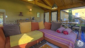 Quarto acolhedor com teto de madeira, cama grande com roupa de cama colorida, mesa de madeira e cadeiras.