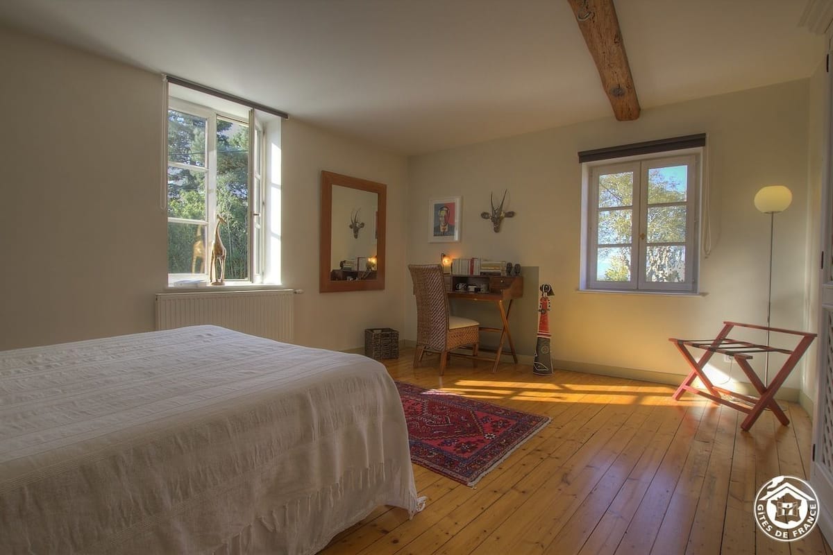 A bedroom with a bed, a desk, and a chair. There are two windows, one with a view of trees and another with a view of the sky.
