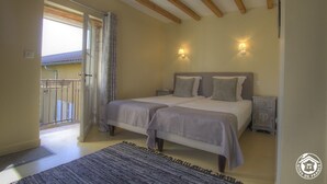 Quarto com duas camas, vigas de madeira no teto, duas luzes fixadas na parede, uma mesinha de cabeceira e vista para a varanda.