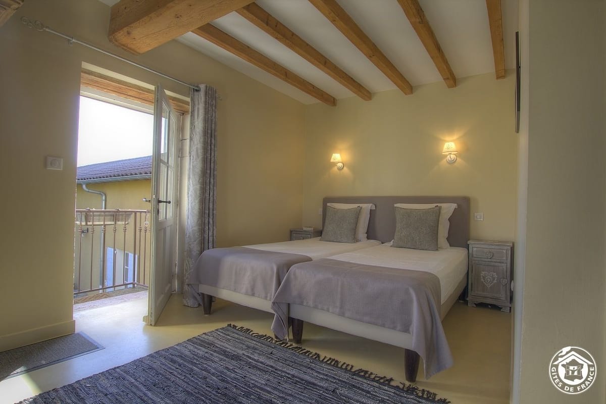 A bedroom with two beds, wooden beams on the ceiling, two wall-mounted lights, a small bedside table, and a view of the outside balcony.