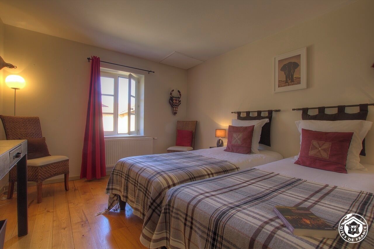 A bedroom with two beds, a desk, a chair, a window with curtains, a wall-mounted lamp, and a framed picture.