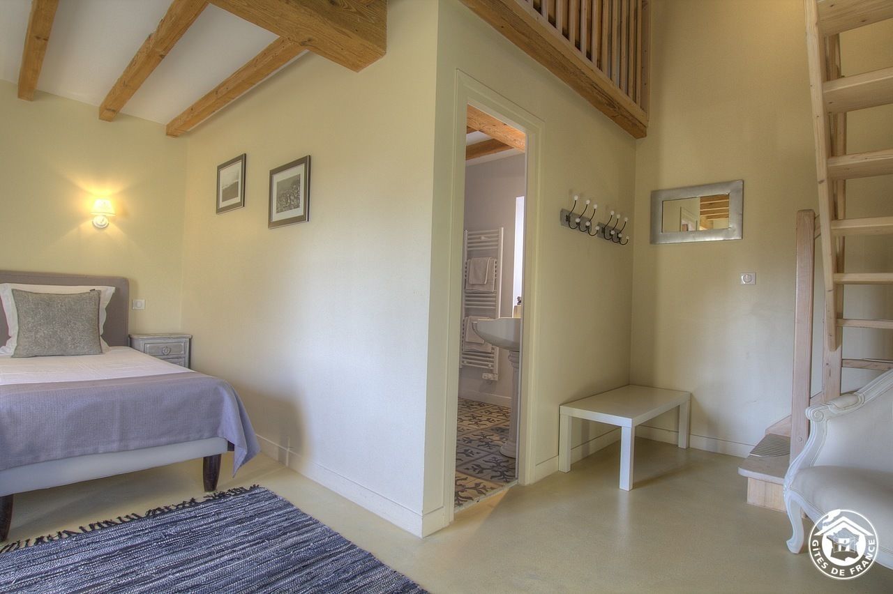A bedroom with a bed, bedside table, and a small bench. There is a door leading to a bathroom with a sink and towel rack.