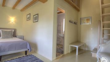 A bedroom with a bed, bedside table, and a small bench. There is a door leading to a bathroom with a sink and towel rack.