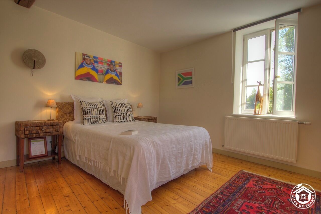 Une chambre avec un grand lit, une fenêtre donnant sur des arbres, un drapeau sud-africain et un tableau coloré accroché au mur.