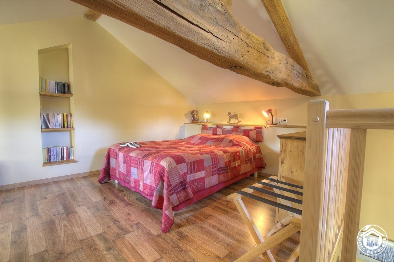 Een slaapkamer met een schuine kap, een houten bed, een nachtkastje met lamp, een boekenplank en een raam.