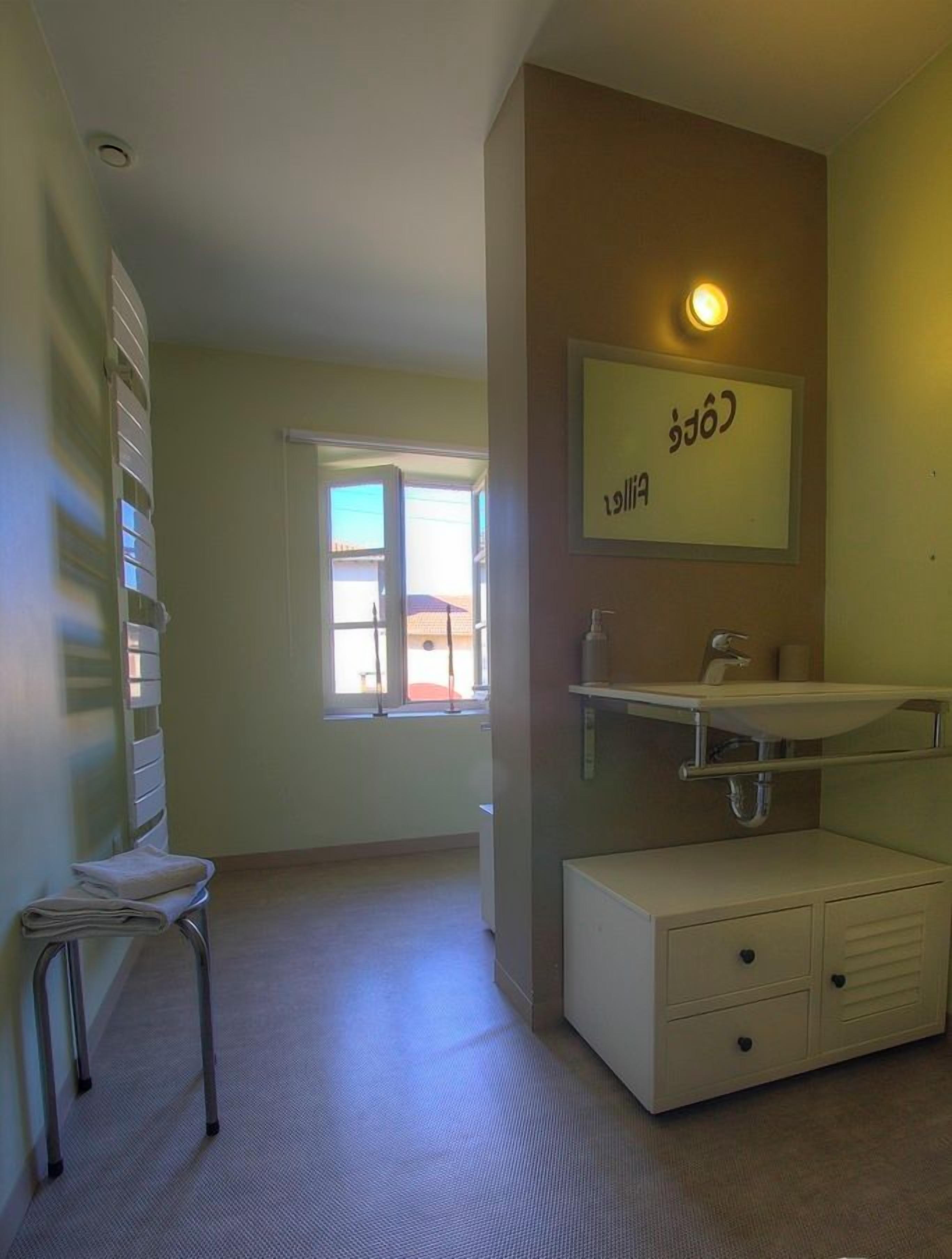 Habitación de hotel con cama, escritorio pequeño, silla, ventana, baño con lavamanos y espejo, y estante montado en la pared.