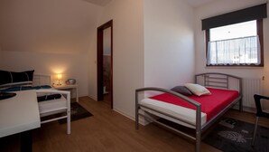 A small, well-lit bedroom with a single bed, a desk with a chair, a window with a view, and a door leading to another room. - Pension Vier Tore (Neubrandenburg)