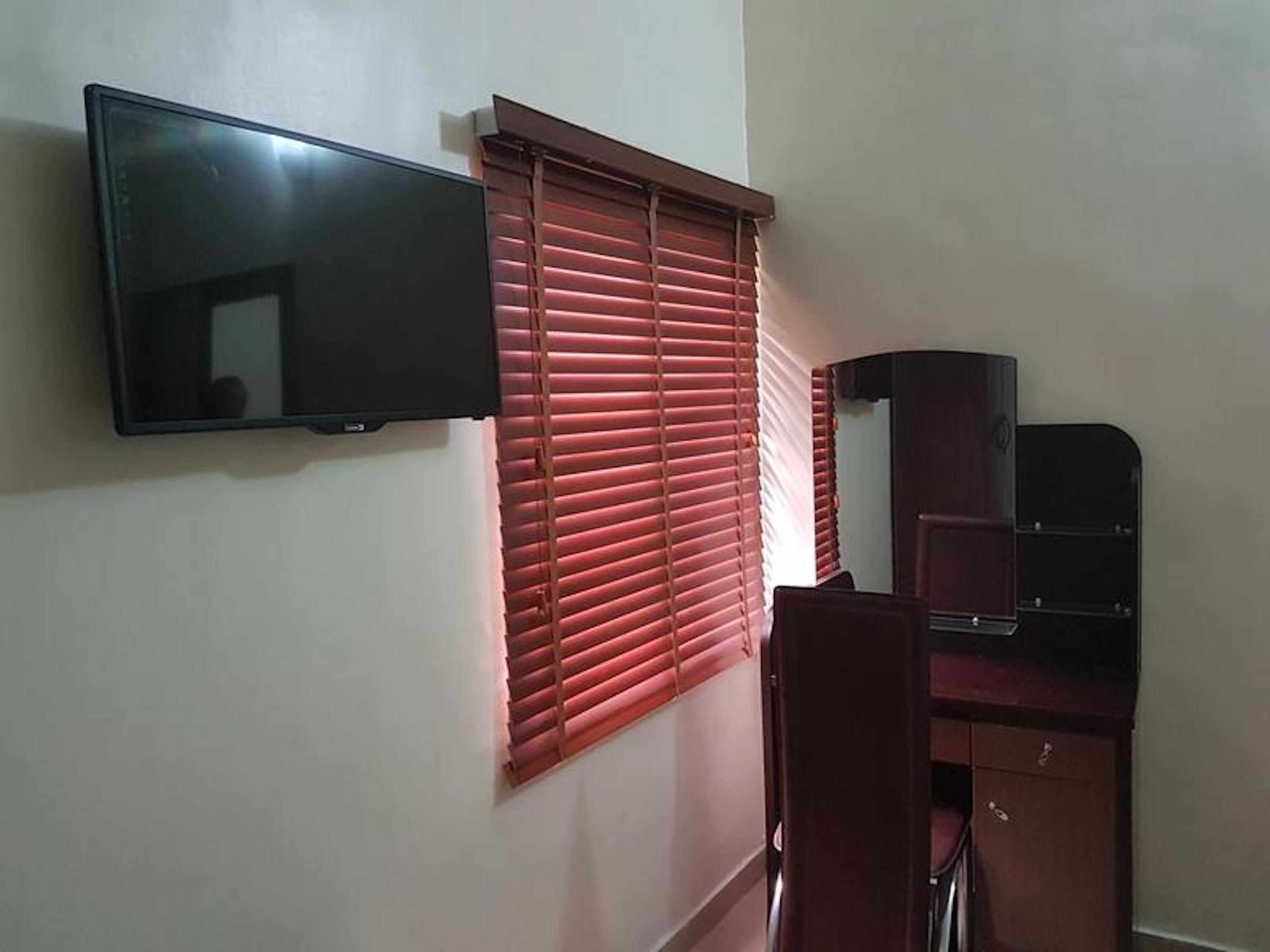 A room with a flat-screen TV mounted on the wall, a window with closed blinds, and a wooden desk with drawers.
