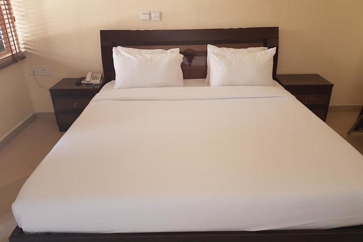 A standard hotel room with a large bed, two bedside tables, a nightstand, and a window with blinds.