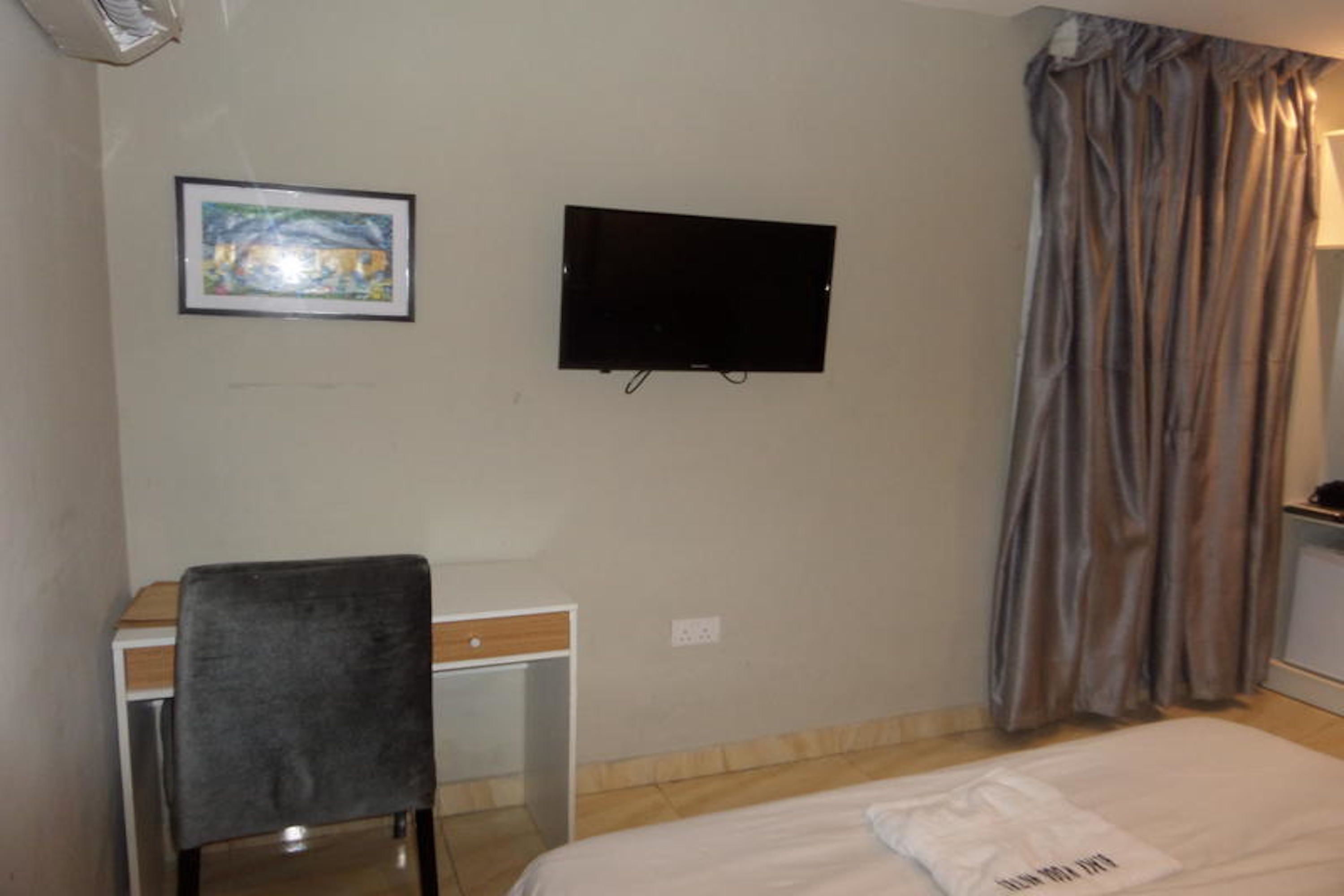 A hotel room with a bed, a desk with a chair, a television mounted on the wall, and a window with curtains.