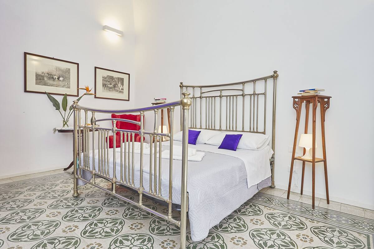 Una habitación con una cama de estructura metálica, una silla roja, una mesita de noche de madera con una lámpara y baldosas estampadas en el suelo.