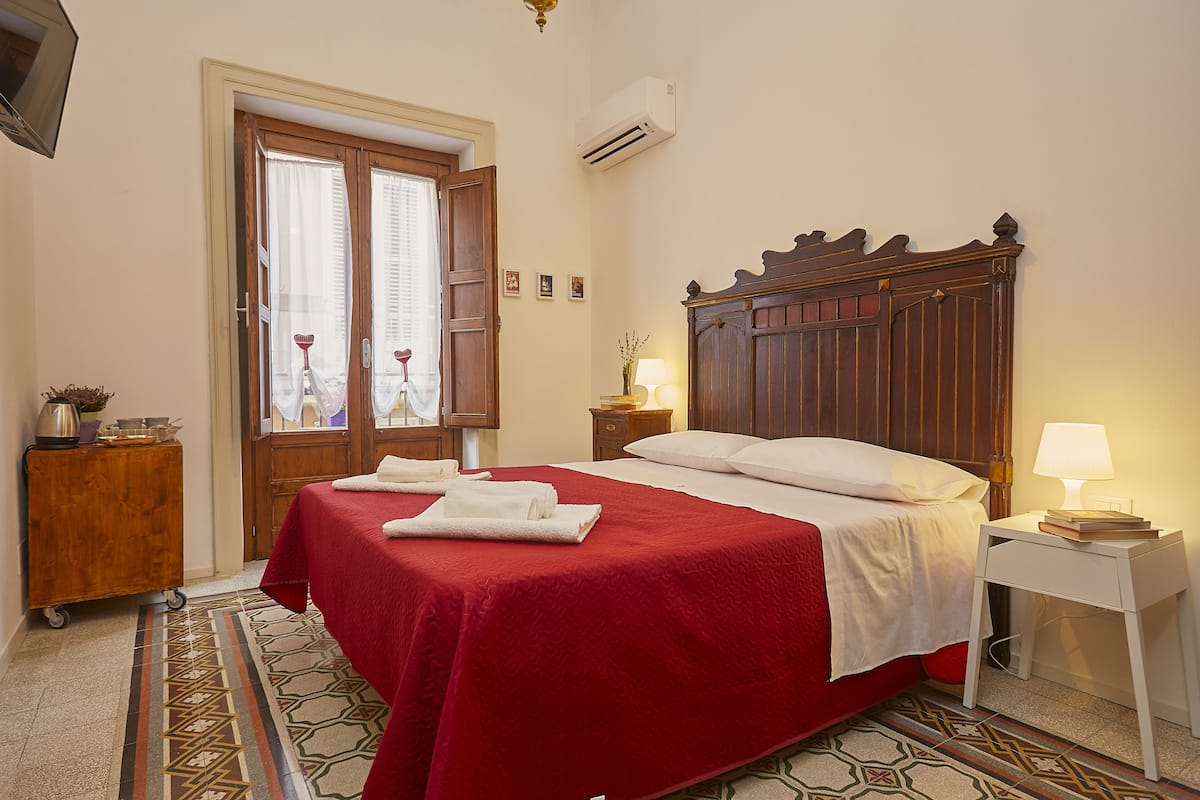 Habitación de hotel con una cama grande, cabecera de madera, mesita de noche con lámpara y ventana con cortinas.