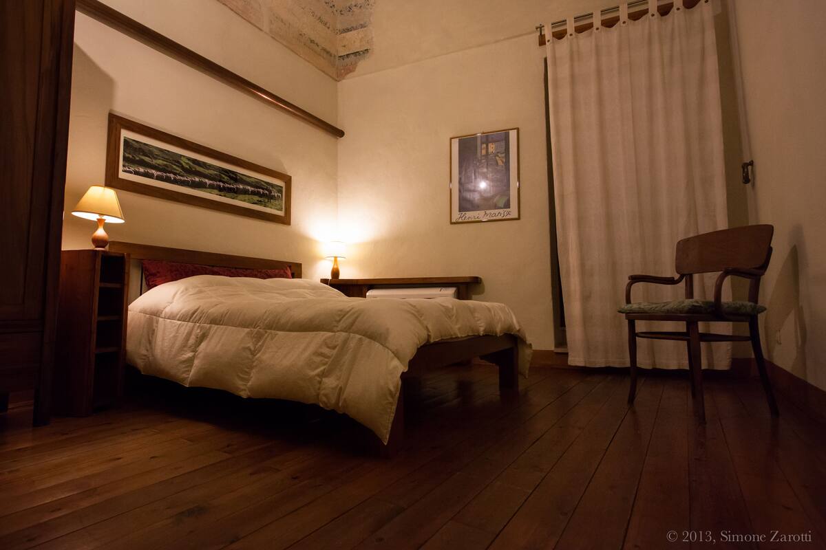 Un dormitorio con cama de madera, una silla, una mesita de noche con lámpara, un cuadro enmarcado en la pared y una ventana con cortinas.