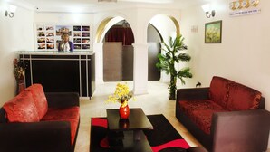 Un vestíbulo de hotel con mostrador de recepción, sofás rojos, una mesa de centro negra con un jarrón de flores y una planta grande en maceta.