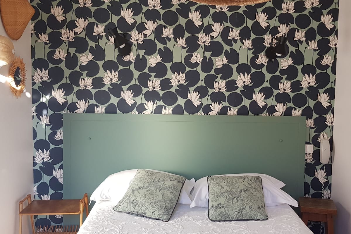 Un lit avec une tête de lit verte, deux oreillers, une table de chevet et un mur recouvert d’un papier peint à motifs floraux.