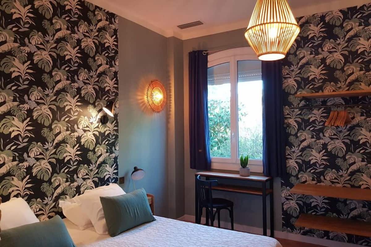 Une chambre à coucher comprenant un lit, un bureau, une chaise, un luminaire suspendu et une fenêtre avec des rideaux.