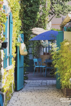 A narrow alleyway with a table and chairs under a blue umbrella, surrounded by greenery and a wicker basket. - Hôtel Les Lauriers (Saint-Tropez)